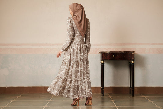 Woman in a long, patterned dress standing in a room with a decorative wall and table.
