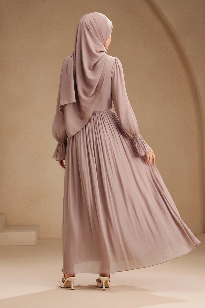 Woman wearing a long, light brown dress against a beige wall.